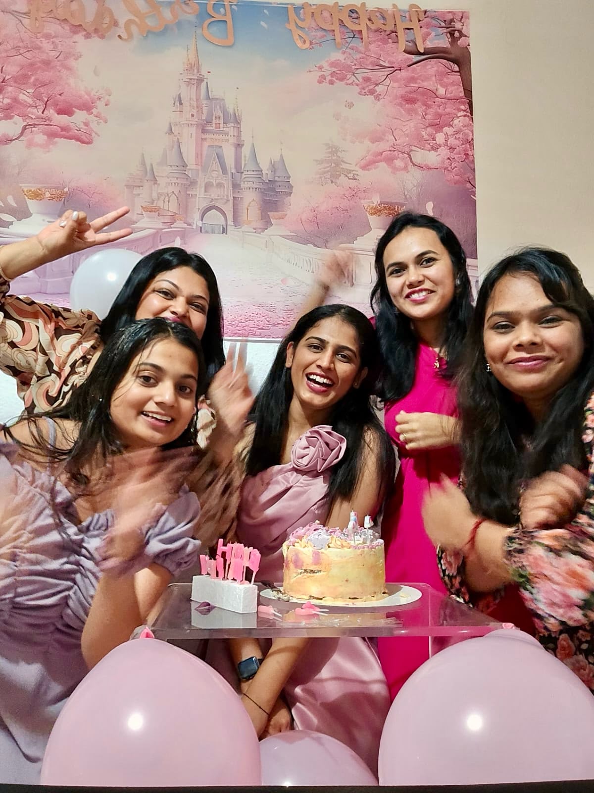 A joyful birthday celebration — friends gathered around a cake with pink balloons and a fairytale-castle backdrop.