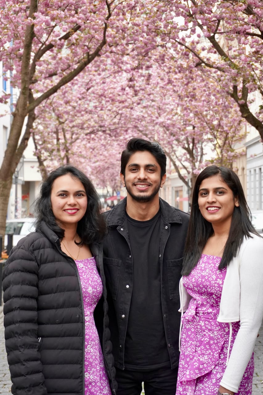 Three friends smiling together under a canopy of pink cherry blossoms on a city street.