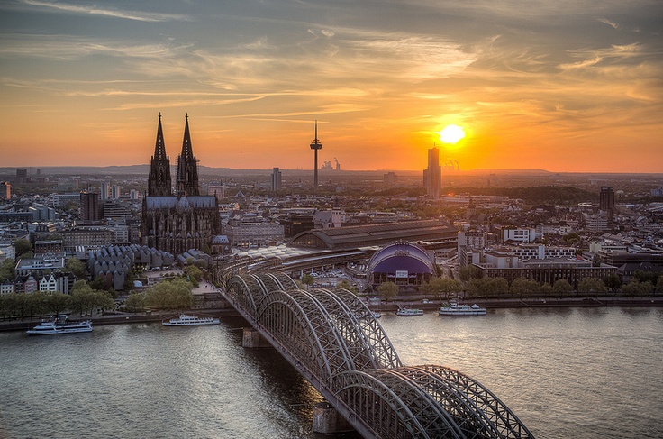 The Cologne skyline at sunset — the cathedral's twin spires, the Hohenzollern Bridge, and the Rhine glowing gold.