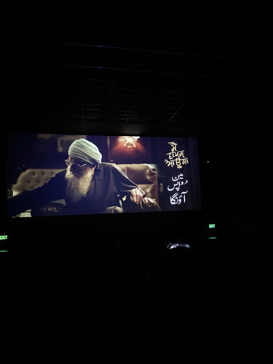 A cinema screen glowing in a dark theatre — an elderly man on screen beside the words 'I will return' in Hindi and Urdu.